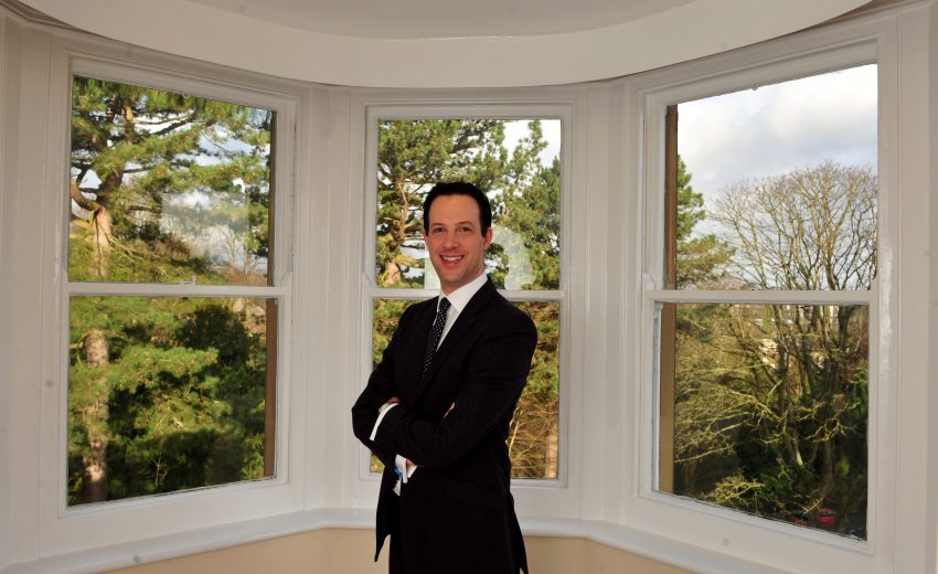 Alex Goldstein stood smiling and with his arms crossed in a large bay window